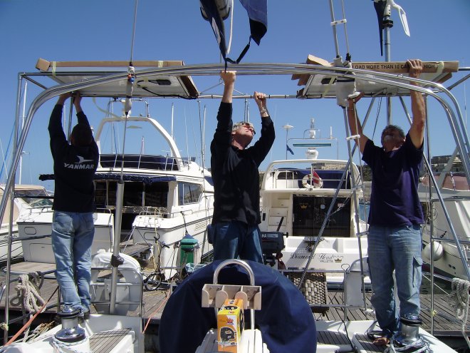 Daniël, Johan en Jaap (vlnr) tillen het frame met de zonnepanelen op zijn plek. De panelen zijn bedekt met karton, ze zijn nog niet aangesloten. In de zon zouden ze de opgewekte stroom niet kwijt kunnen en beschadigd raken. Rechtsboven de windgenerator