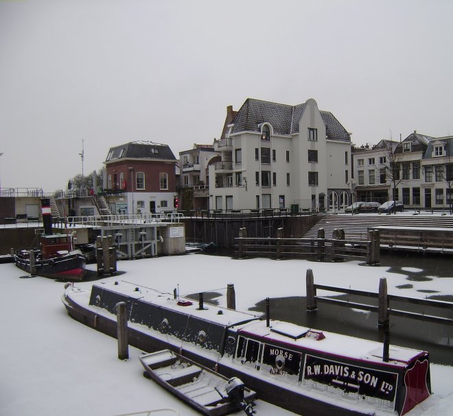 In het witte appartementengebouw aan de Lingehaven kunnen we een appartement huren op de begane grond (waar de rolgordijnen van dicht zijn) Links het sluisje naar de Merwede en de sleepboot `Jan de Sterke`