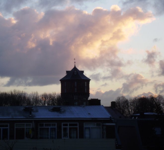 De zonsopgang tovert goud op de wolken. Ervoor het zwarte silhouet van de watertoren. Dat alles voor het slaapkamerraam vanmorgen
