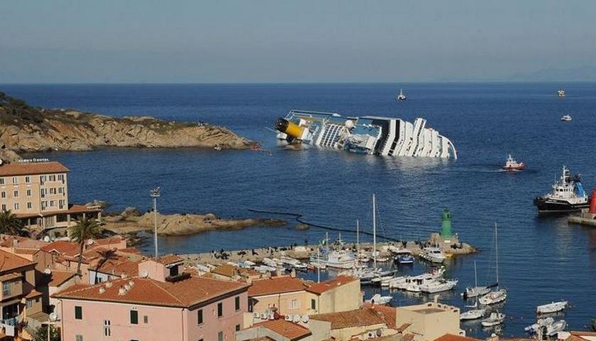 De Costa Concordia op zijn zij bij het haventje van Giglio (rechtsonder)